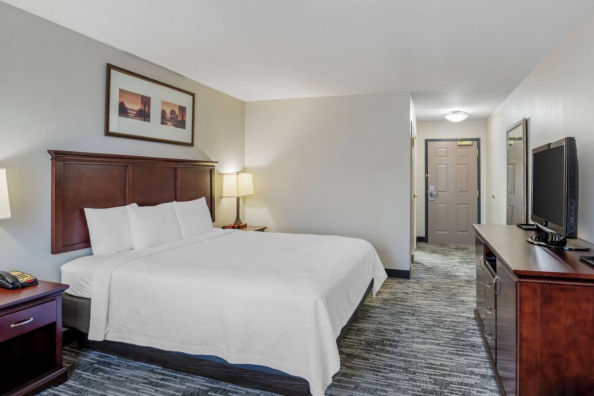 A hotel room with a large bed, bedside lamps, a wooden headboard, a flat-screen TV, a desk with a telephone, and a carpeted floor.