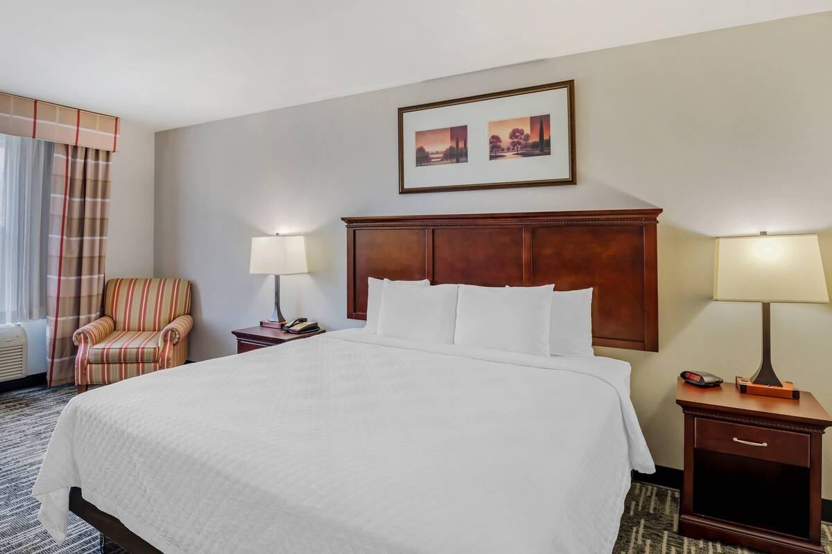 A hotel room with a large bed, a wooden headboard, two bedside lamps, a chair, a nightstand, and a framed picture on the wall.