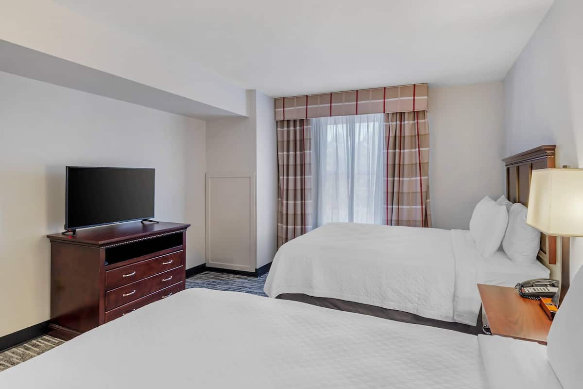 A hotel room with a flat-screen TV, a wooden dresser, two beds, a nightstand with a lamp, and a window with curtains.