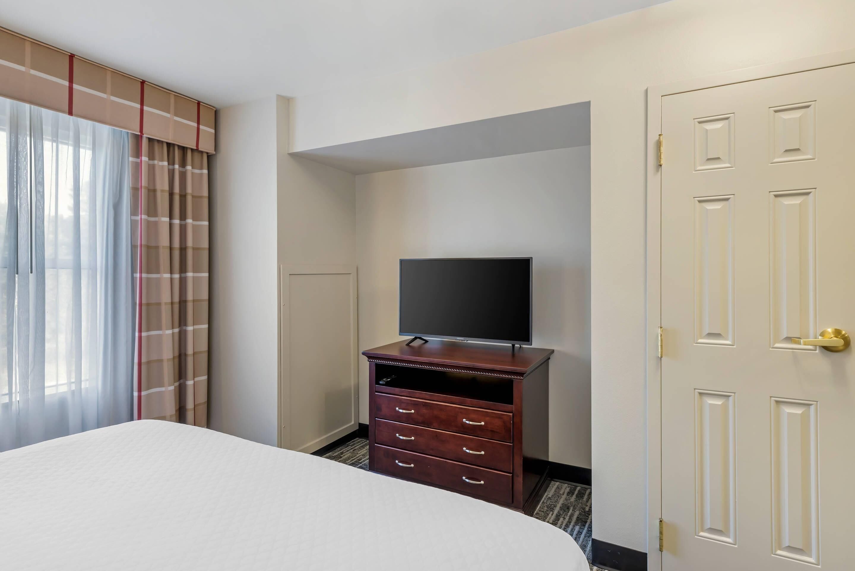 A hotel room with a bed, a TV on a wooden dresser, a door, and a window with curtains.