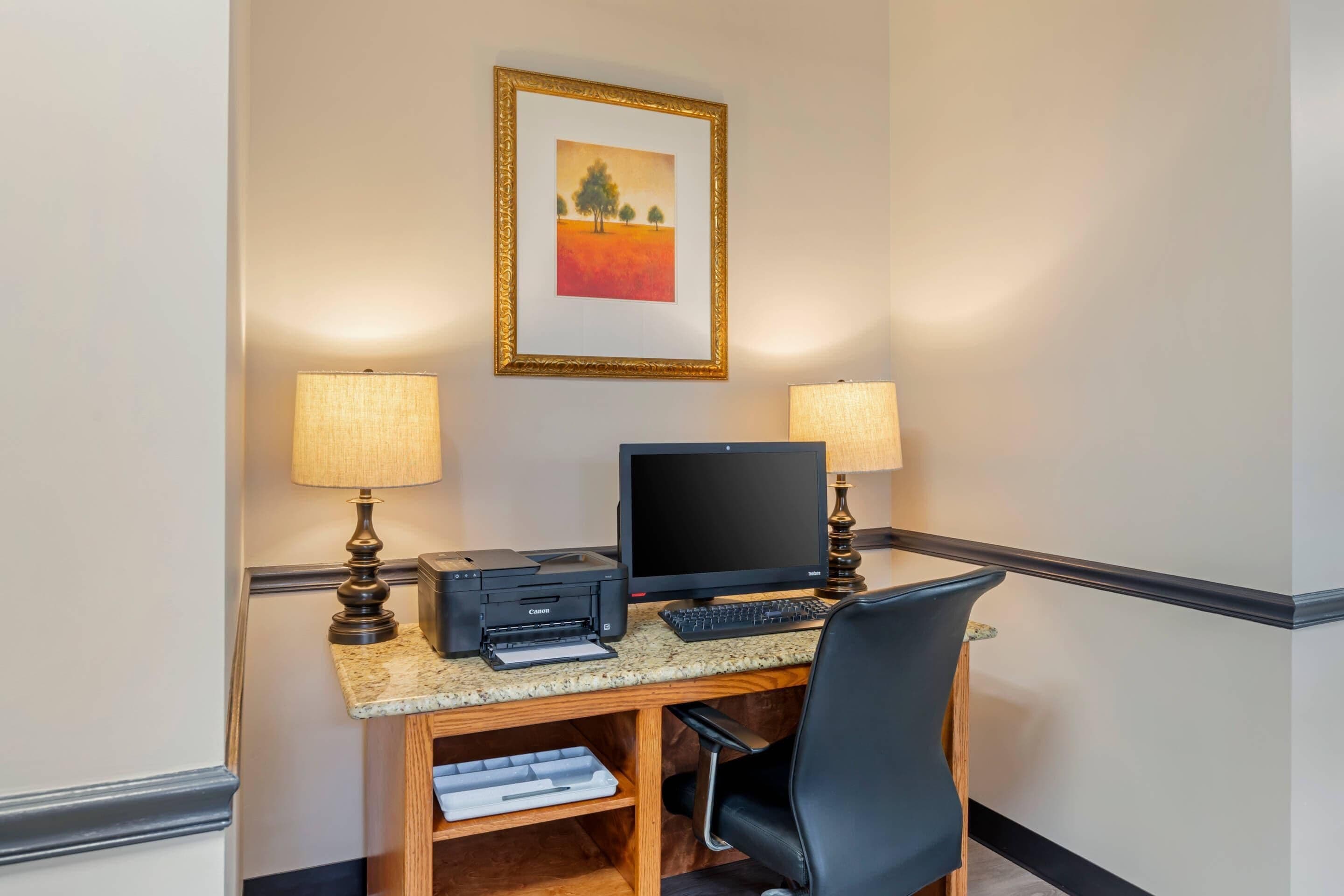 A hotel room with a desk, two lamps, a framed painting, and a printer.