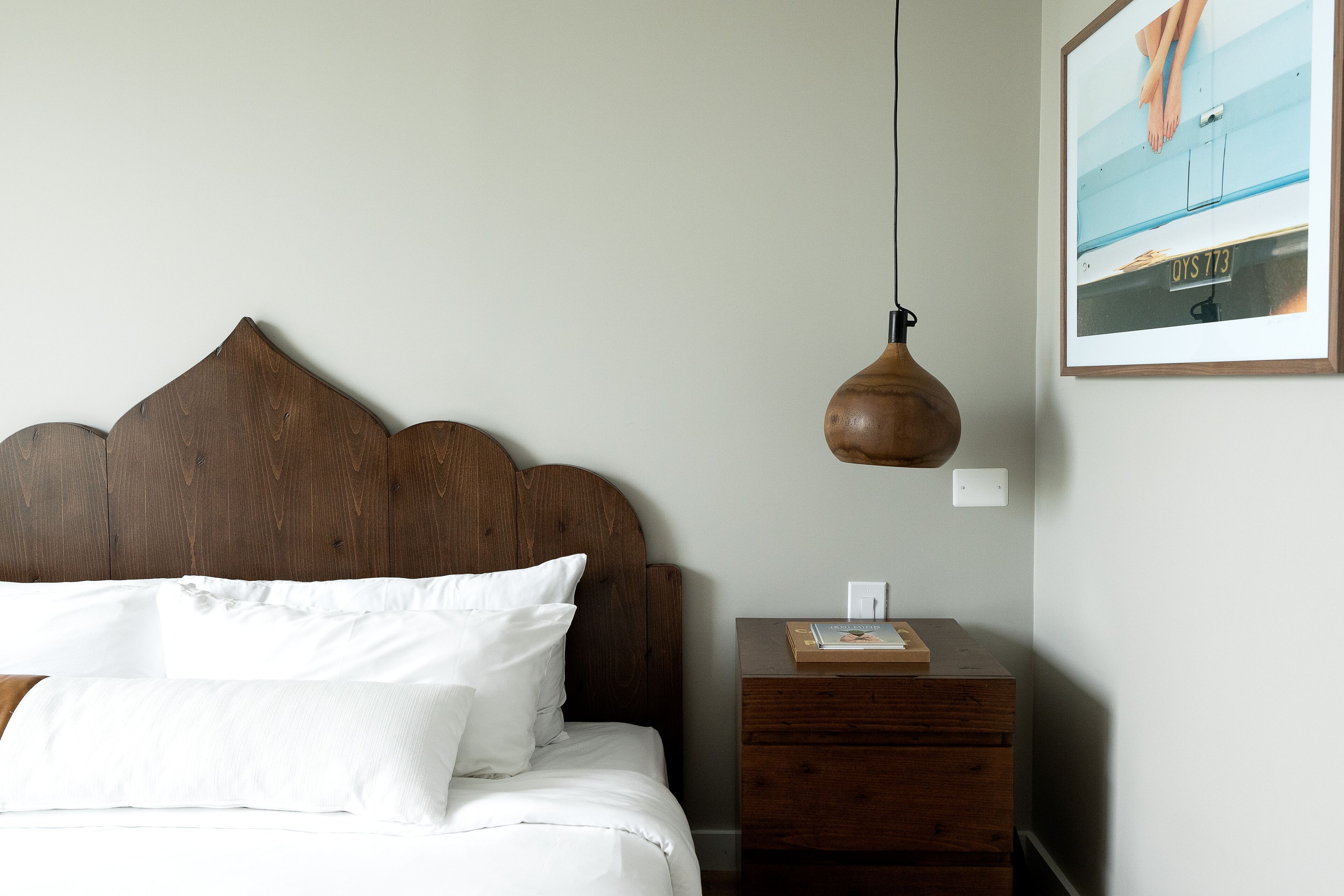 Un lit bien fait, avec du linge de lit blanc et une tête de lit en bois, une petite table de chevet en bois sur laquelle se trouvent un livre et une lampe, et un tableau encadré accroché au mur.