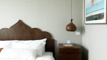 Un lit bien fait, avec du linge de lit blanc et une tête de lit en bois, une petite table de chevet en bois sur laquelle se trouvent un livre et une lampe, et un tableau encadré accroché au mur.