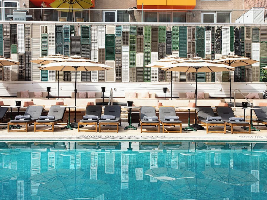 A poolside area with lounge chairs and umbrellas, a tiled pool, and a building with shutters in the background.