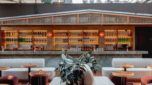 Zona de bar con sofás y mesas redondas, gran variedad de botellas en estanterías y vistas a edificios al fondo.