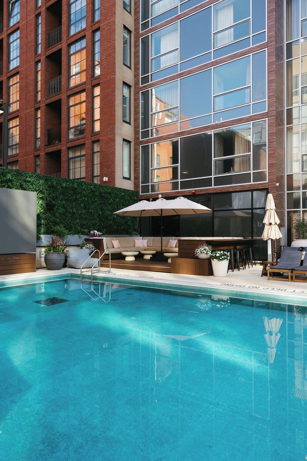 Piscina con sillas de descanso y sombrillas, rodeada por un edificio de ladrillo con ventanas amplias.