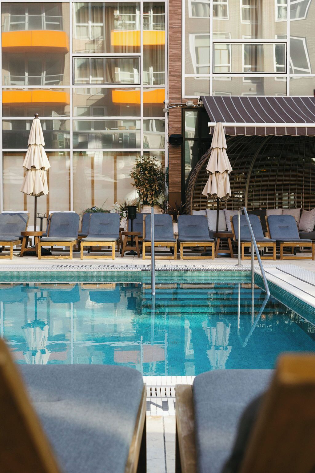 Ein Poolbereich mit Liegestühlen und Sonnenschirmen, ein gefliester Pool und ein Gebäude mit großen Fenstern und Balkonen.