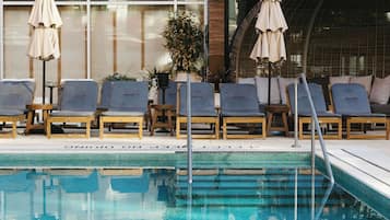 Ein Poolbereich mit Liegestühlen und Sonnenschirmen, ein gefliester Pool und ein Gebäude mit großen Fenstern und Balkonen.