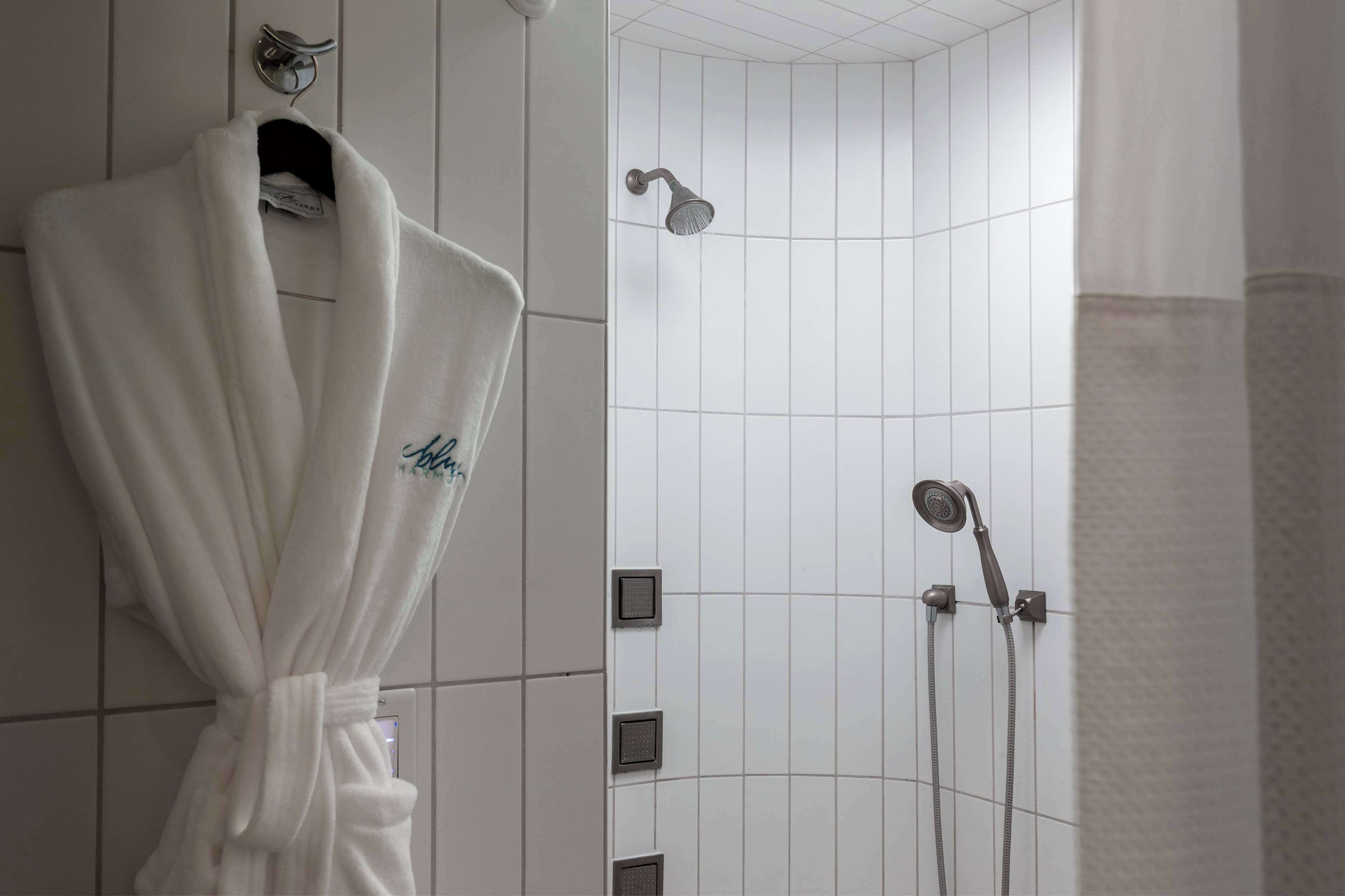 A bathroom with a white tiled wall, a white robe hanging on a hook, and a shower with two showerheads.