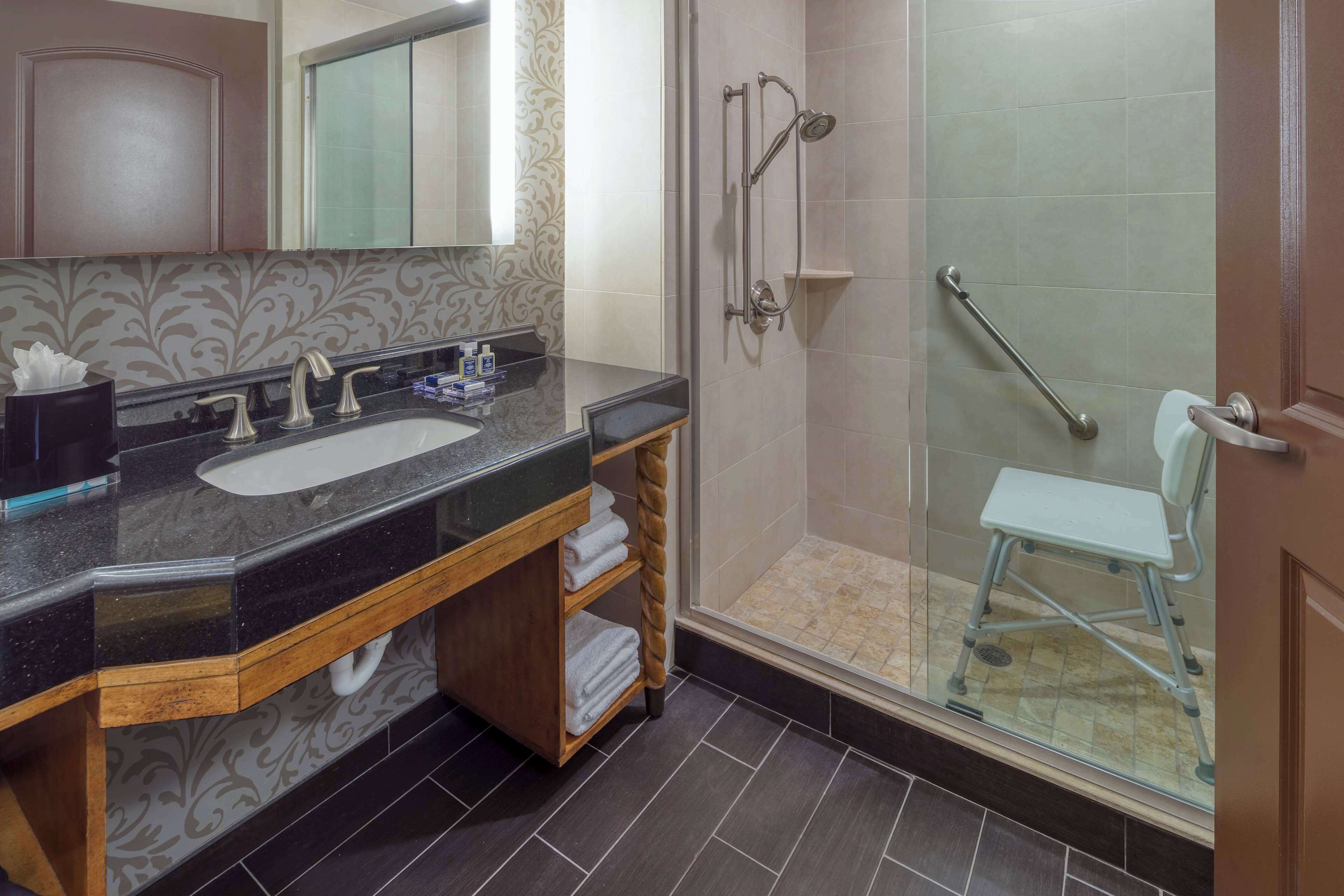 A bathroom with a black countertop sink, a mirror, a shower with a glass door, and a white chair.
