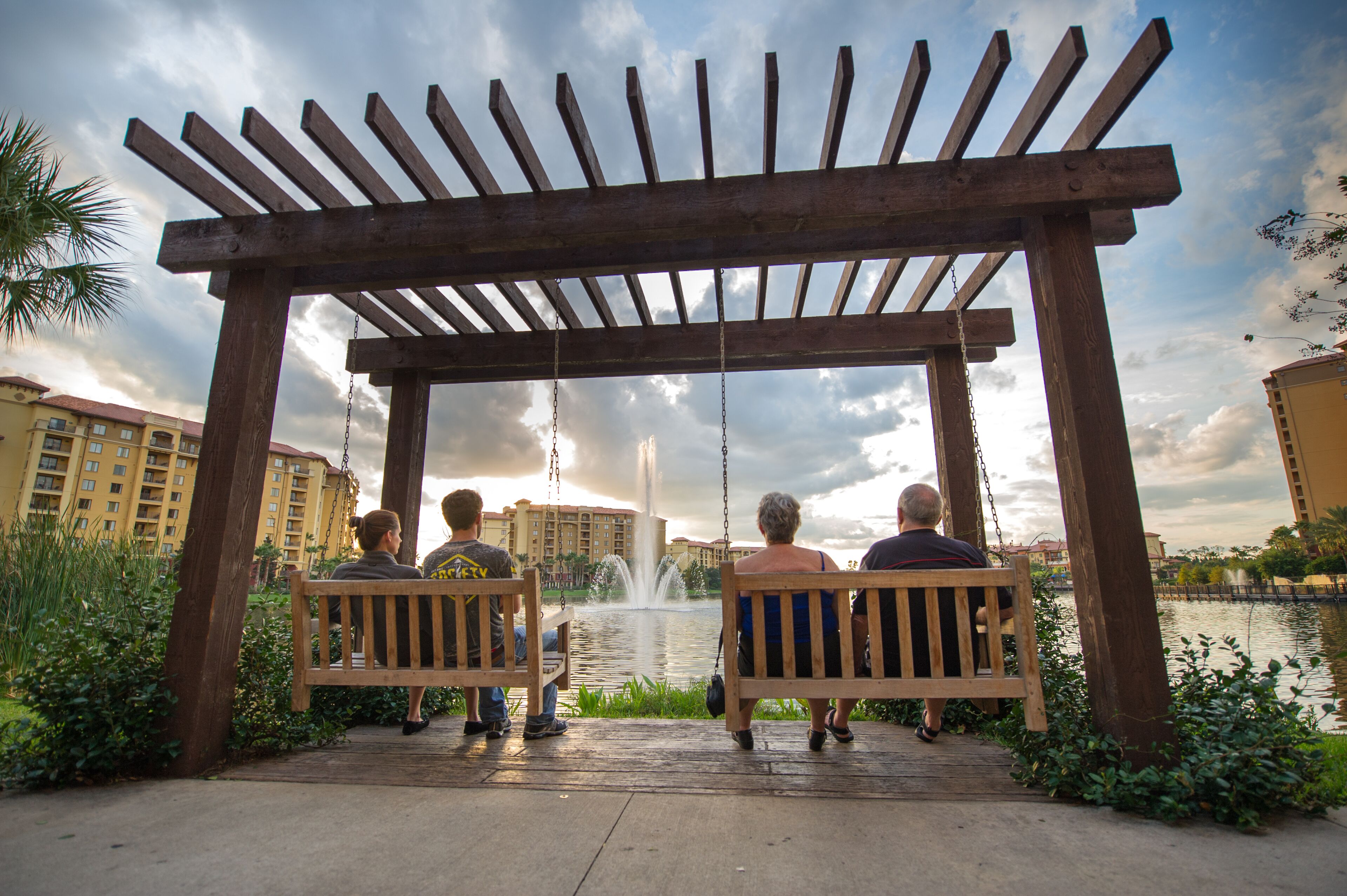 Een houten pergola met schommels, met uitzicht op een waterpartij en gebouwen op de achtergrond.