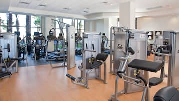 A gym with various exercise machines, large windows, and a wooden floor.