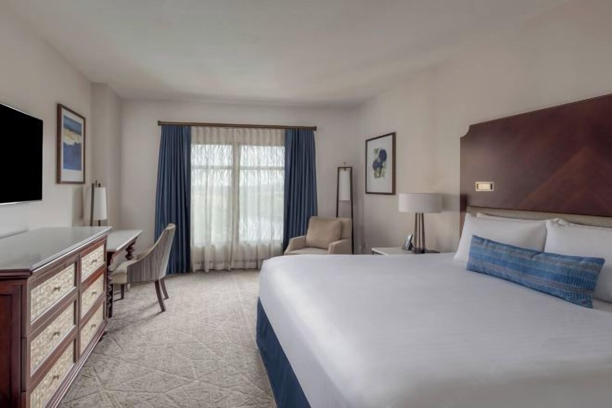 Habitación de hotel con una cama grande, un escritorio, una silla, un televisor y una ventana con cortinas.