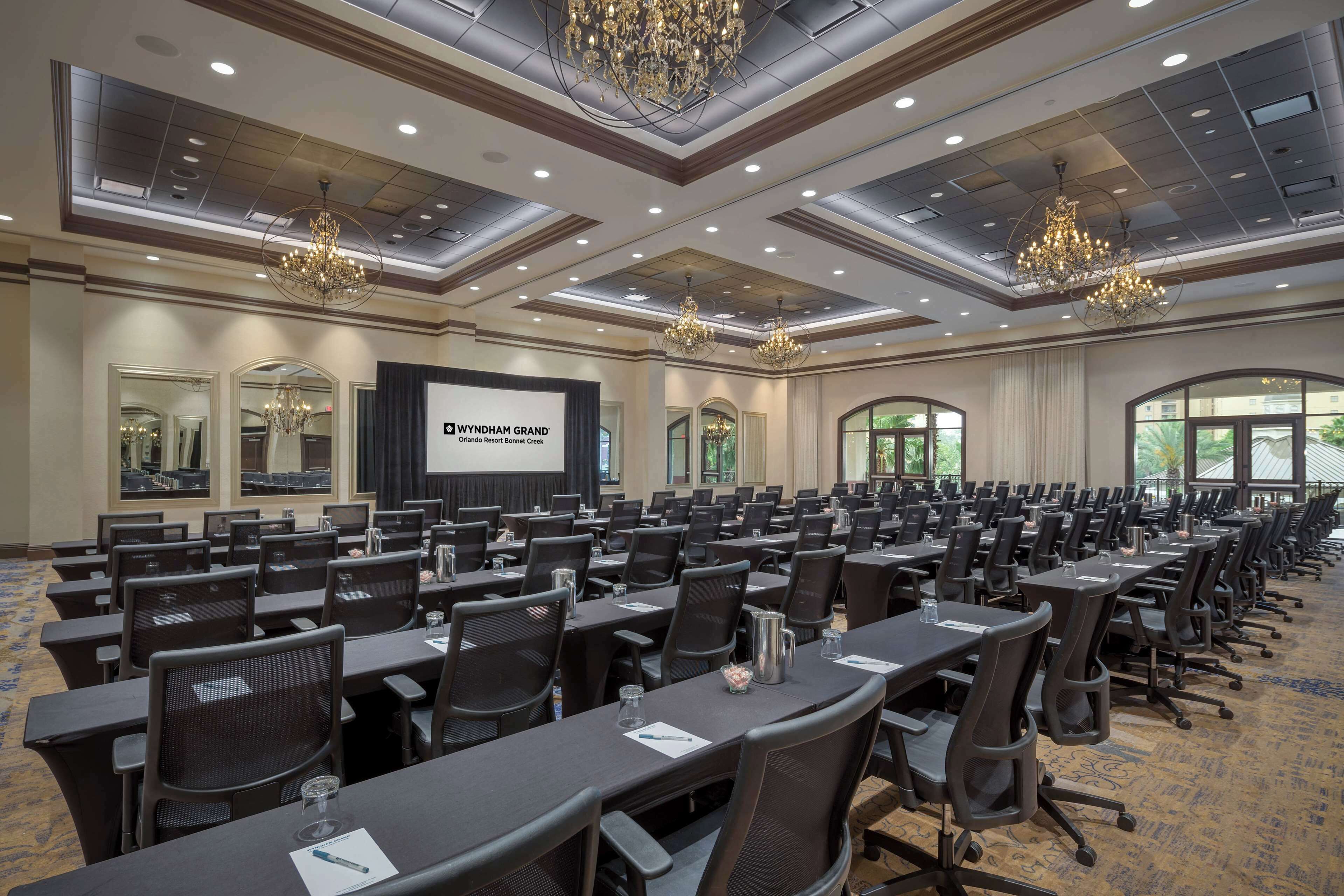 A conference room with rows of tables and chairs, chandeliers, and a projection screen displaying "WYNDHAM GRAND".