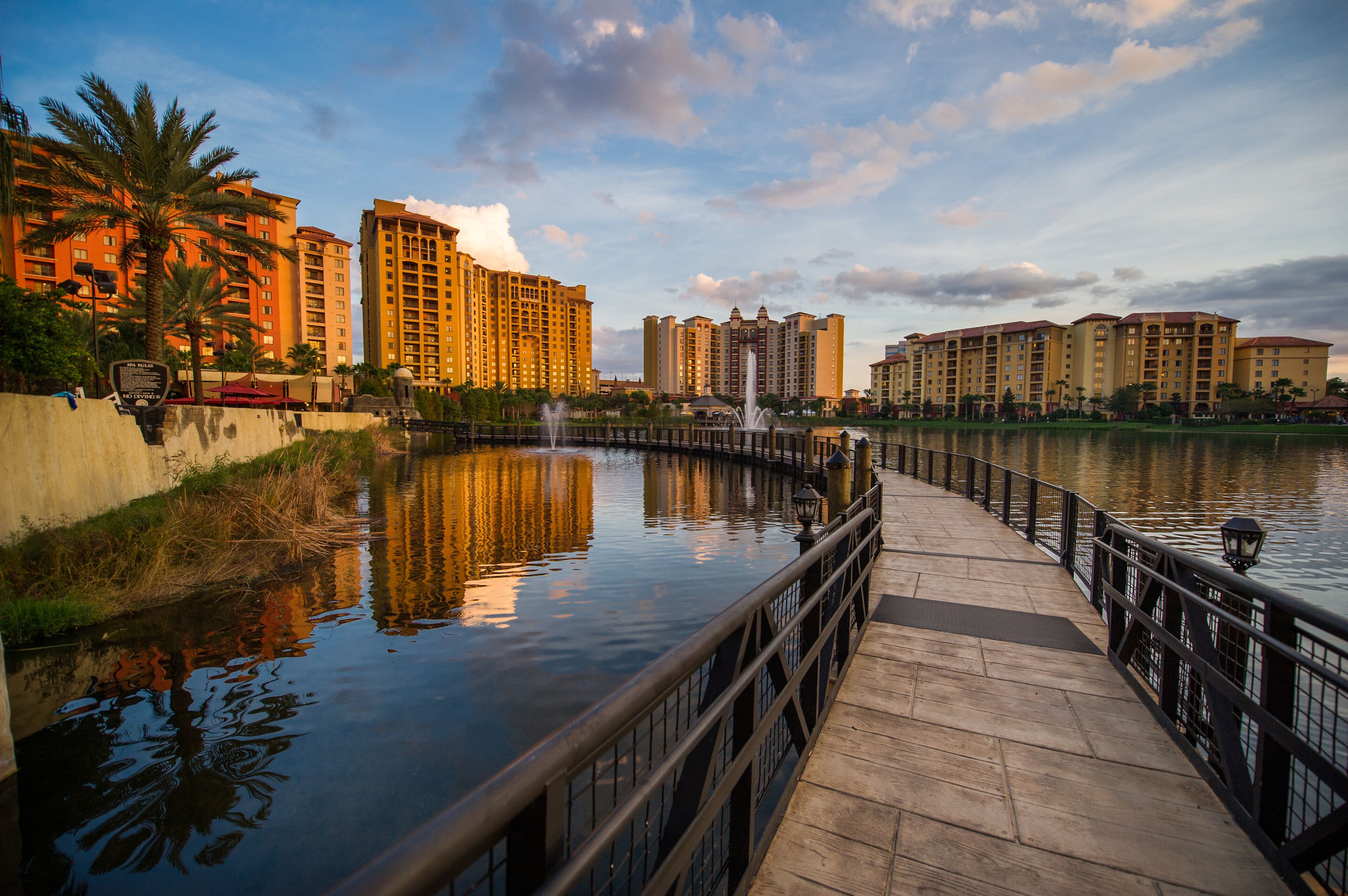 A waterfront promenade with a reflective water surface, a wooden walkway, and high-rise buildings in the background.