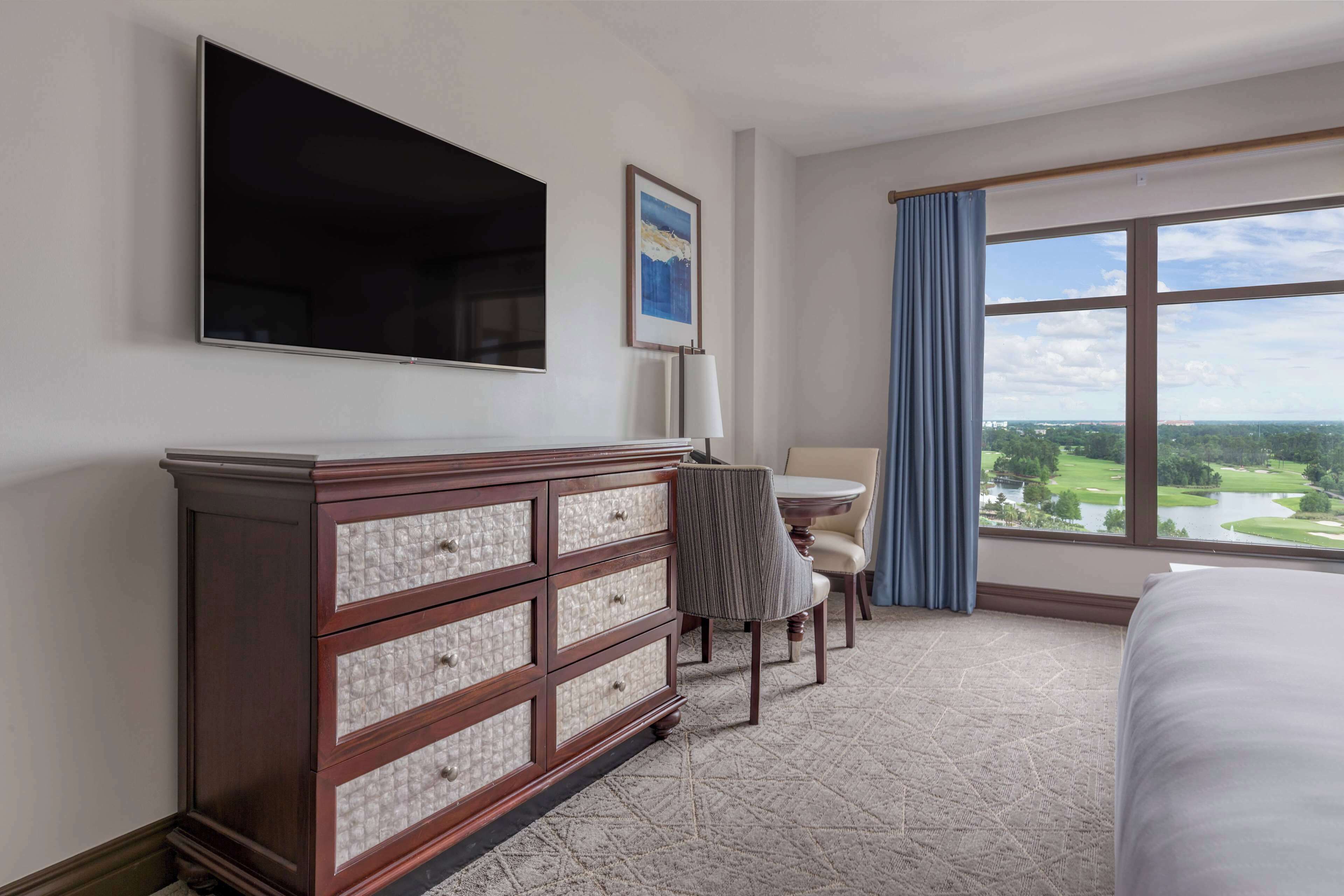 A hotel room with a large bed, a dresser, a TV mounted on the wall, a dining table with chairs, and a large window with a view of a golf course.