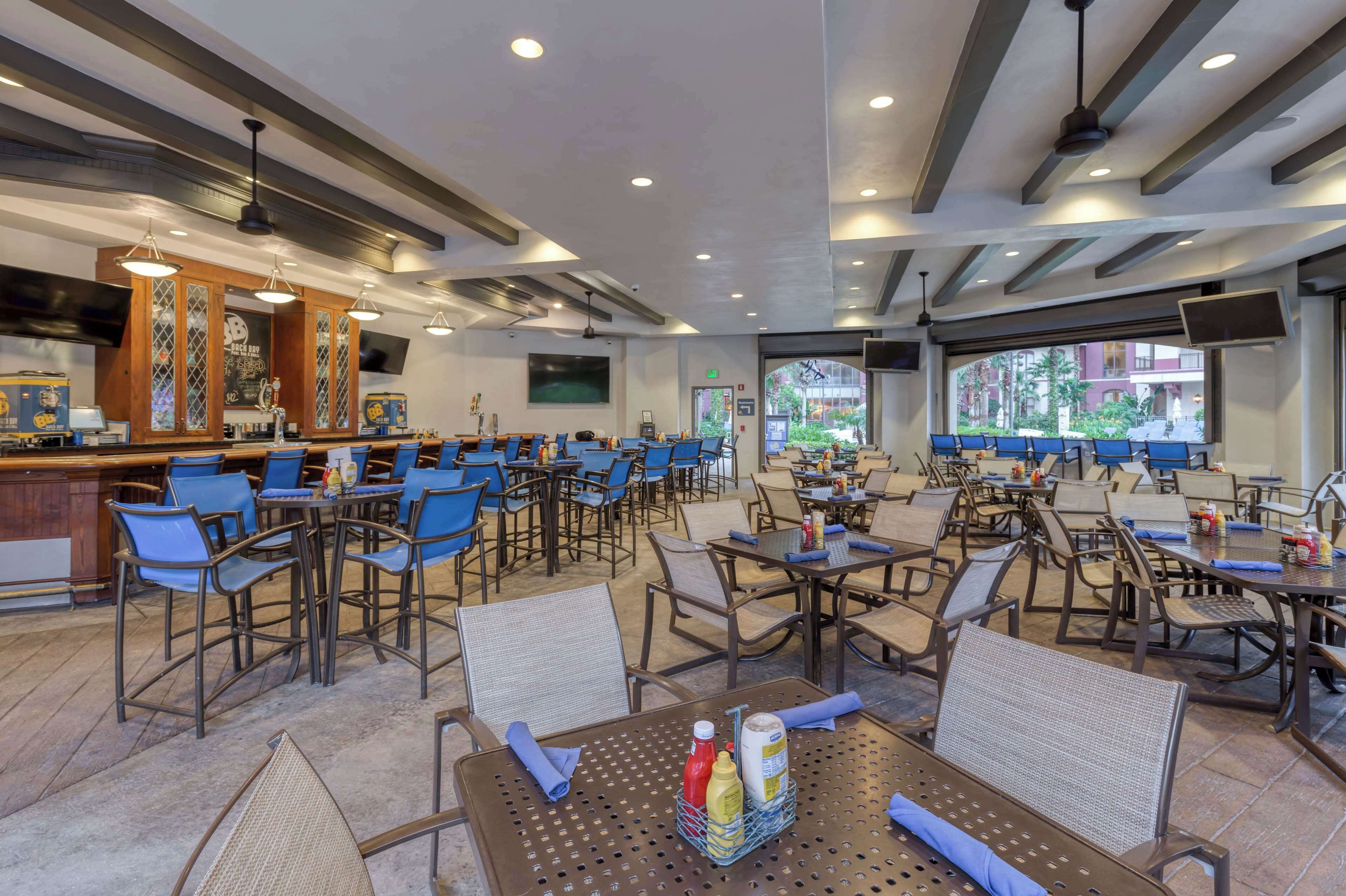 Un amplio comedor interior con varias mesas y sillas, una zona de bar con mostrador, y grandes ventanales que ofrecen vistas al exterior.