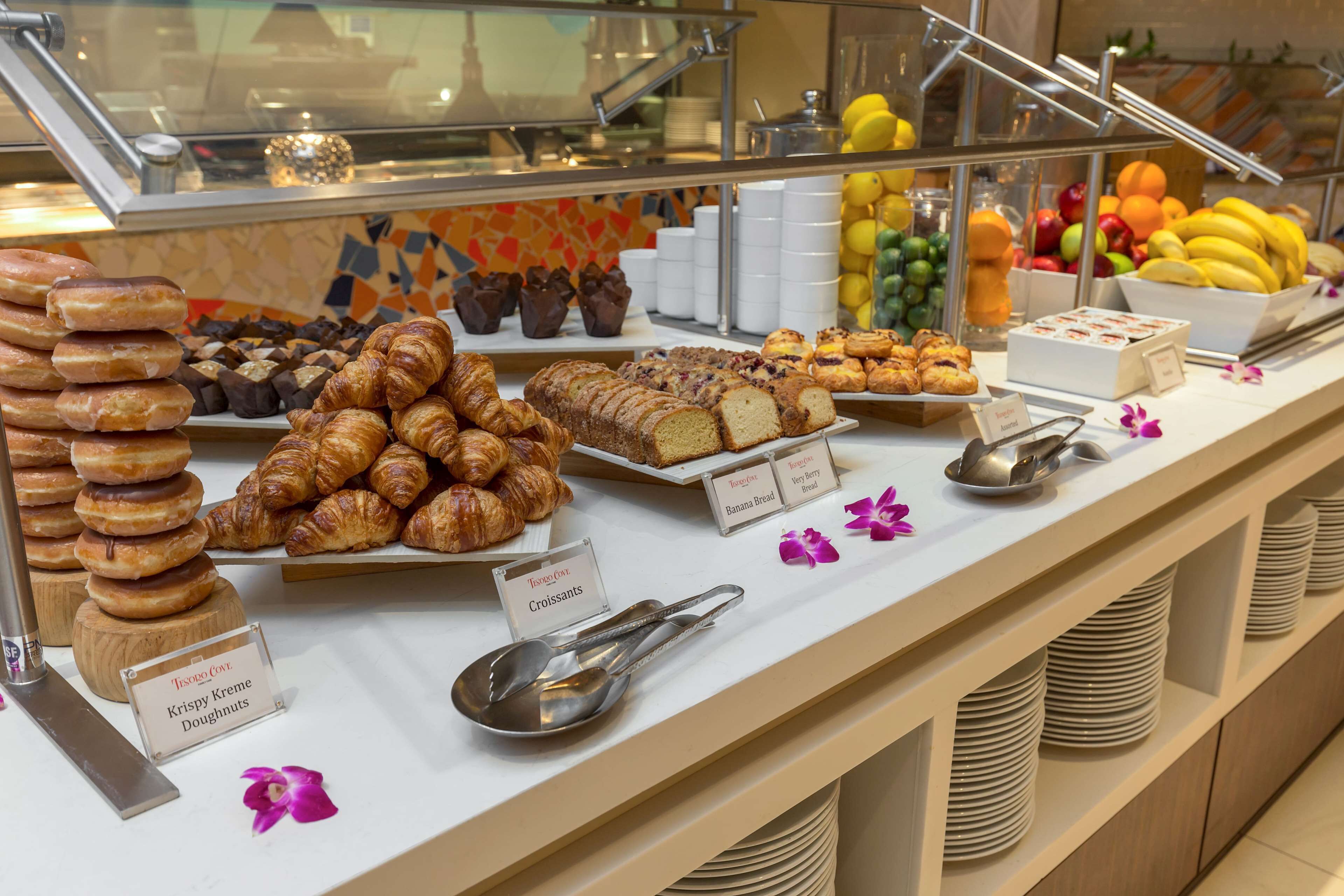 A buffet with various baked goods like croissants, donuts, and muffins, along with fresh fruits and labeled serving dishes.