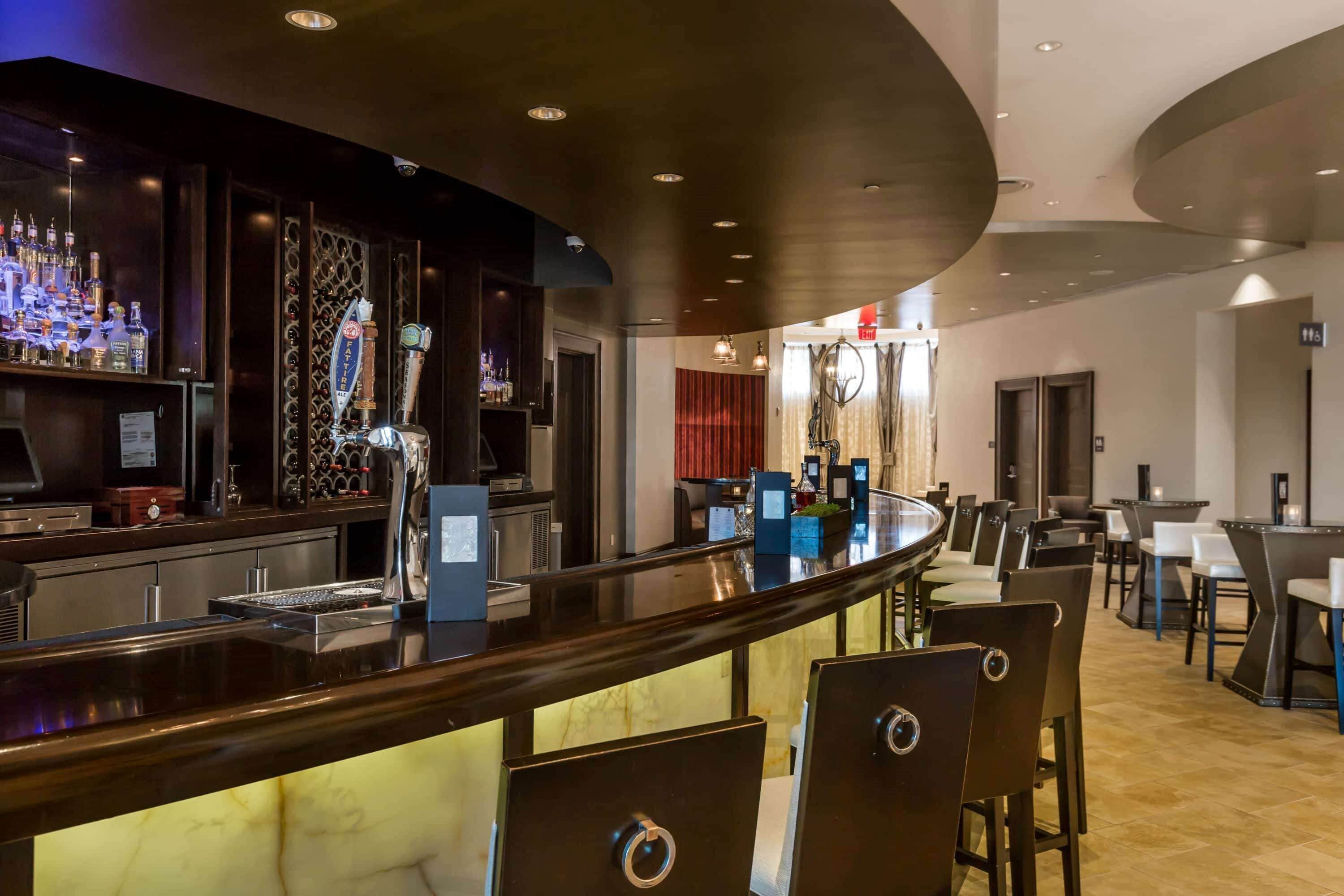 A bar area with a curved counter, bar stools, and a selection of bottles. There are tables and chairs in the adjacent dining area.