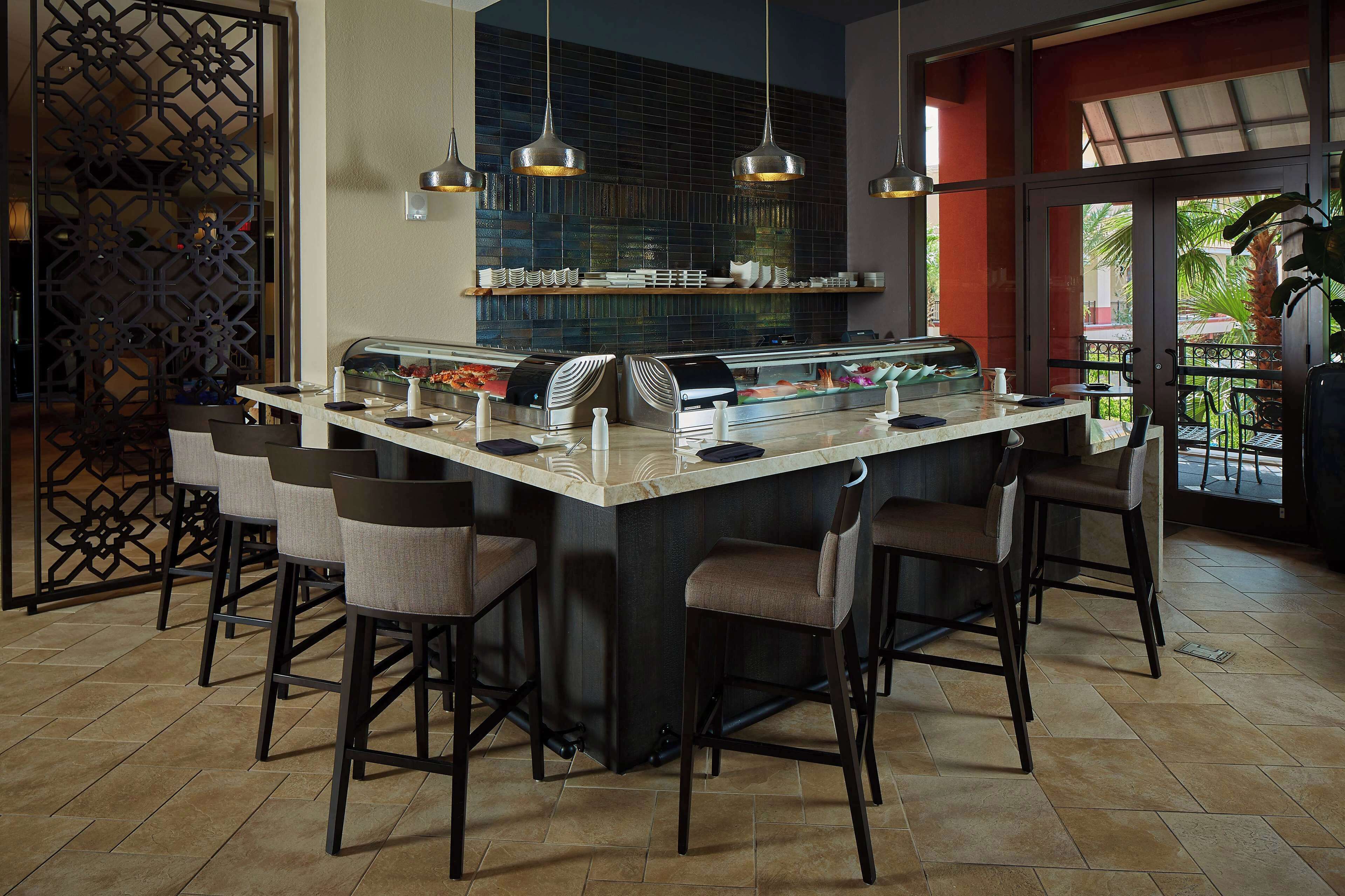 Een bar met een centraal aanrecht, verschillende barkrukken, hangende吊灯 en uitzicht op het terras via grote glazen deuren.