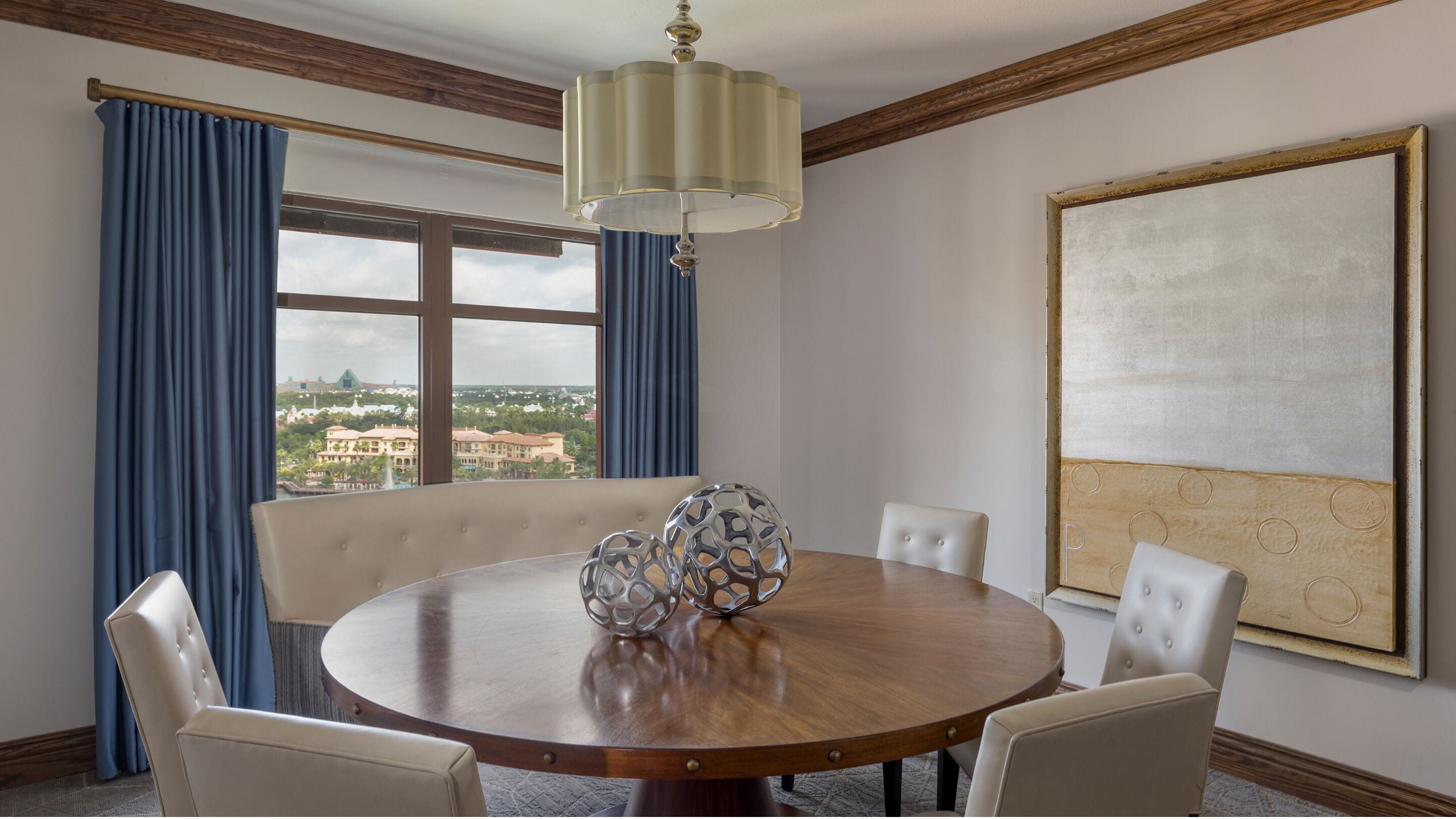 Una mesa redonda de comedor con acabado de madera, rodeada por cuatro sillas tapizadas. Una escultura decorativa de metal está sobre la mesa. La habitación cuenta con un ventanal con cortinas azules, un candelabro y una obra de arte grande enmarcada en la pared.