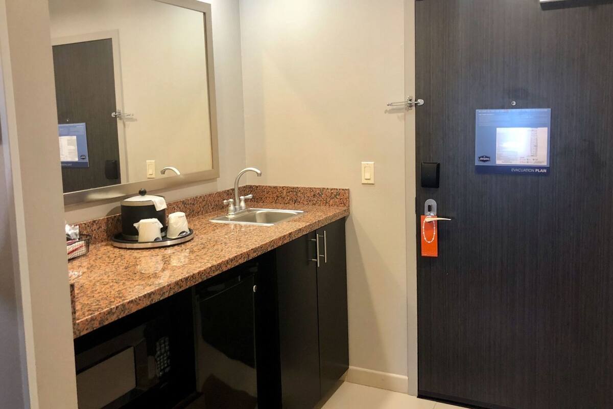 A modern bathroom with a granite countertop, a sink, a mirror, and a dark door with an emergency exit sign.
