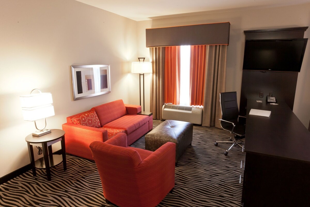 A hotel room with a sofa, ottoman, a desk with a chair, a TV, a lamp, and a large window with curtains.
