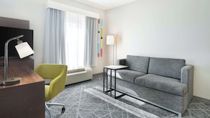 A modern hotel room with a grey sofa, a yellow chair, a desk with a lamp, a flat-screen TV, and a large window with curtains.