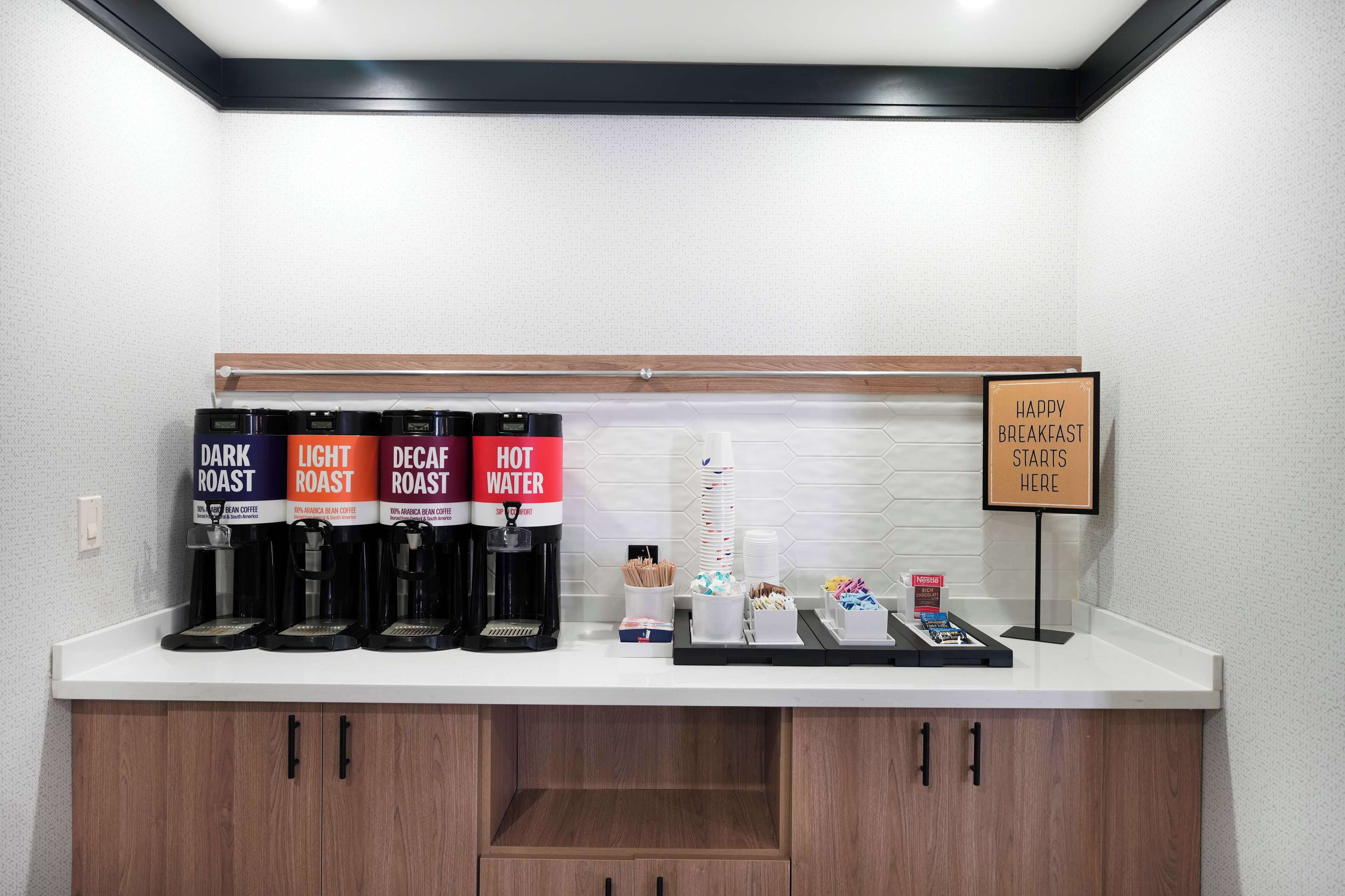 A coffee station with four coffee dispensers labeled DARK ROAST, LIGHT ROAST, DECAF ROAST, and HOT WATER. There are also stacks of disposable cups and a sign that says HAPPY BREAKFAST STARTS HERE.
