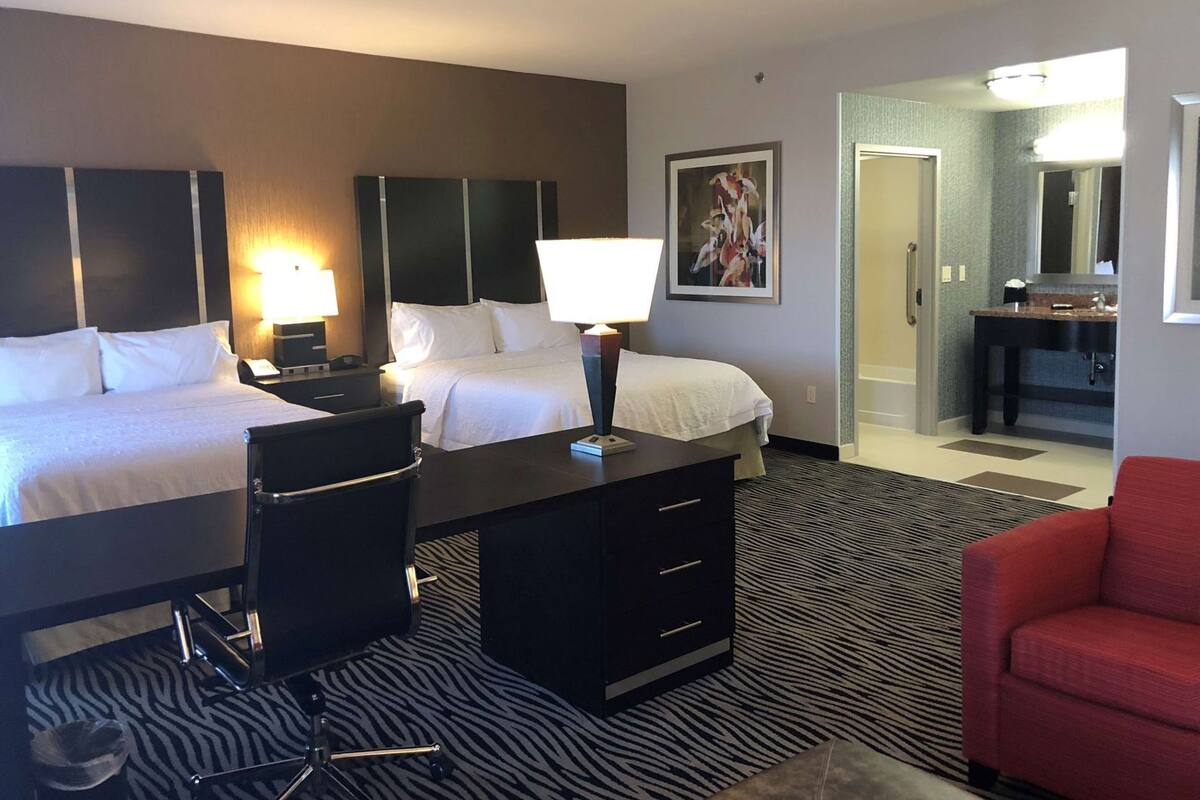 A hotel room with two beds, a desk, a chair, a red sofa, a painting on the wall, and a bathroom visible through an open door.