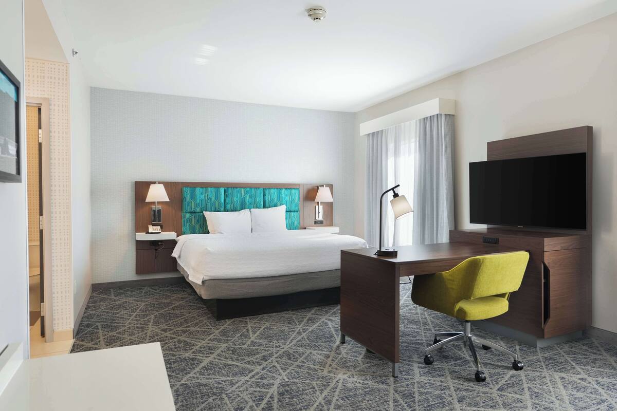 A hotel room with a bed, desk, chair, TV, lamps, and a bathroom door.