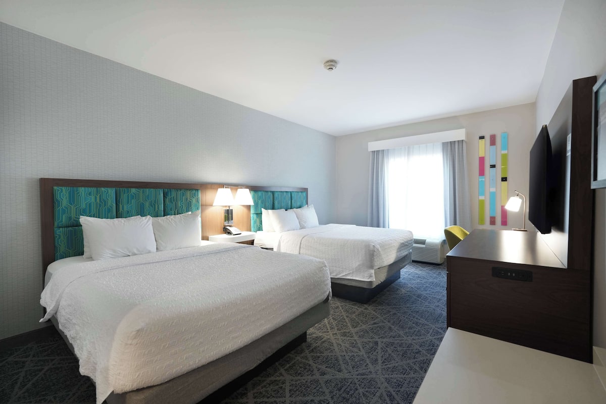 A hotel room with two beds, a desk, a TV, and a window with curtains.