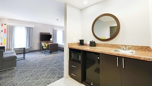 A modern hotel room with a kitchenette, a large mirror, a flat-screen TV, a gray sofa, and a patterned carpet.