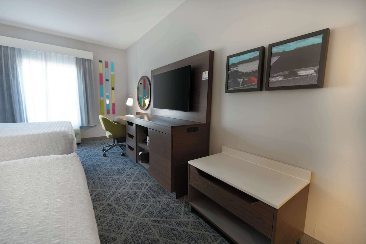 A hotel room with two beds, a desk with a flat-screen TV, a chair, and artwork on the wall.