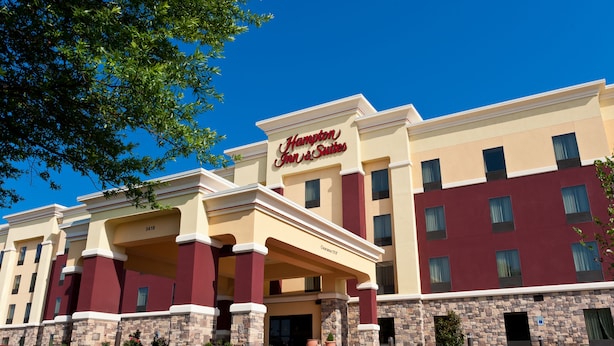 A Hampton Inn & Suites building with a beige and maroon facade, stone accents, and a clear blue sky.