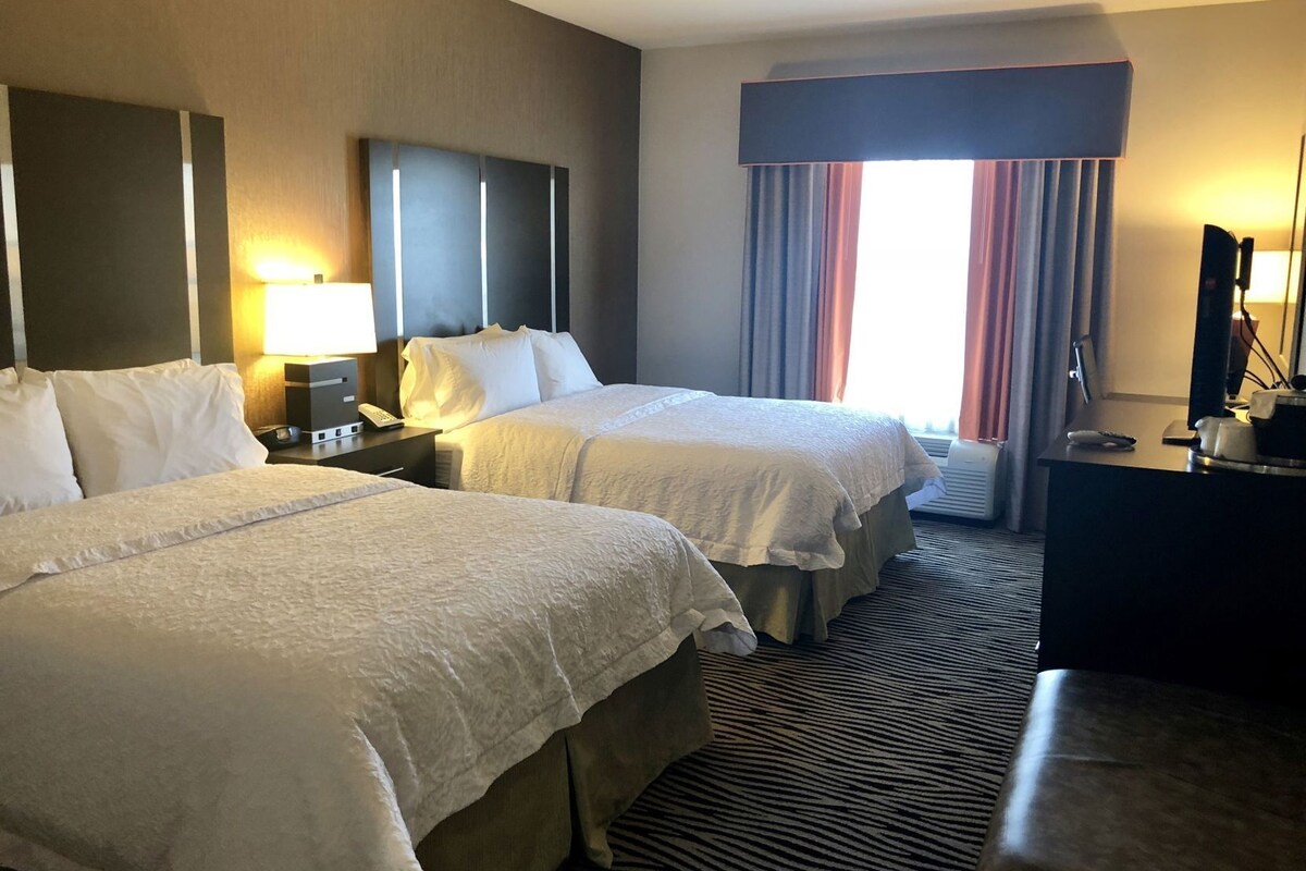 A hotel room with two beds, a desk, a lamp, and a large window with curtains.