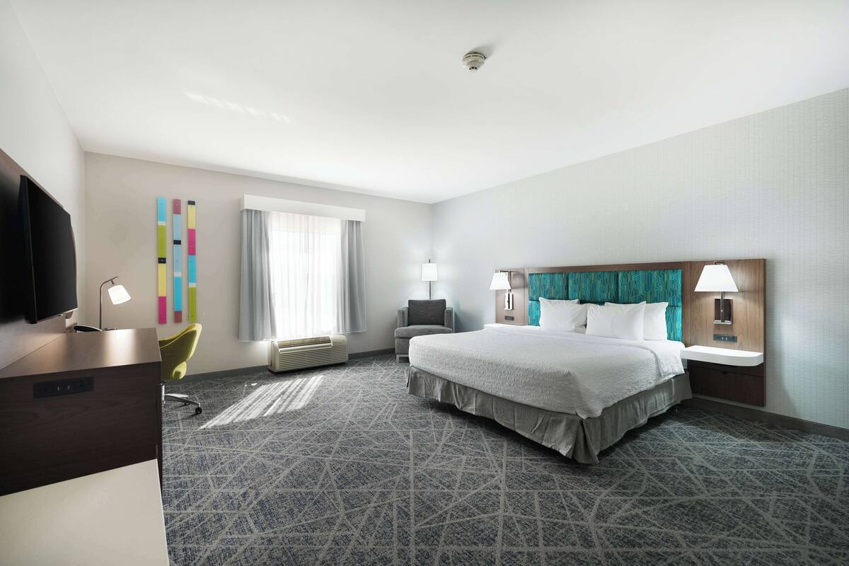 A hotel room with a large bed, a desk with a computer, a chair, a TV, and a window with curtains.
