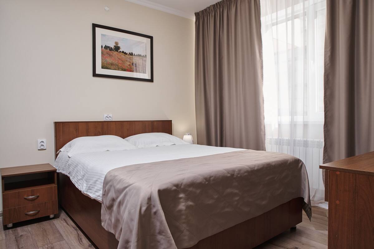 A neatly made bed with a wooden headboard, a bedside table with drawers, a framed picture on the wall, and a window with curtains.