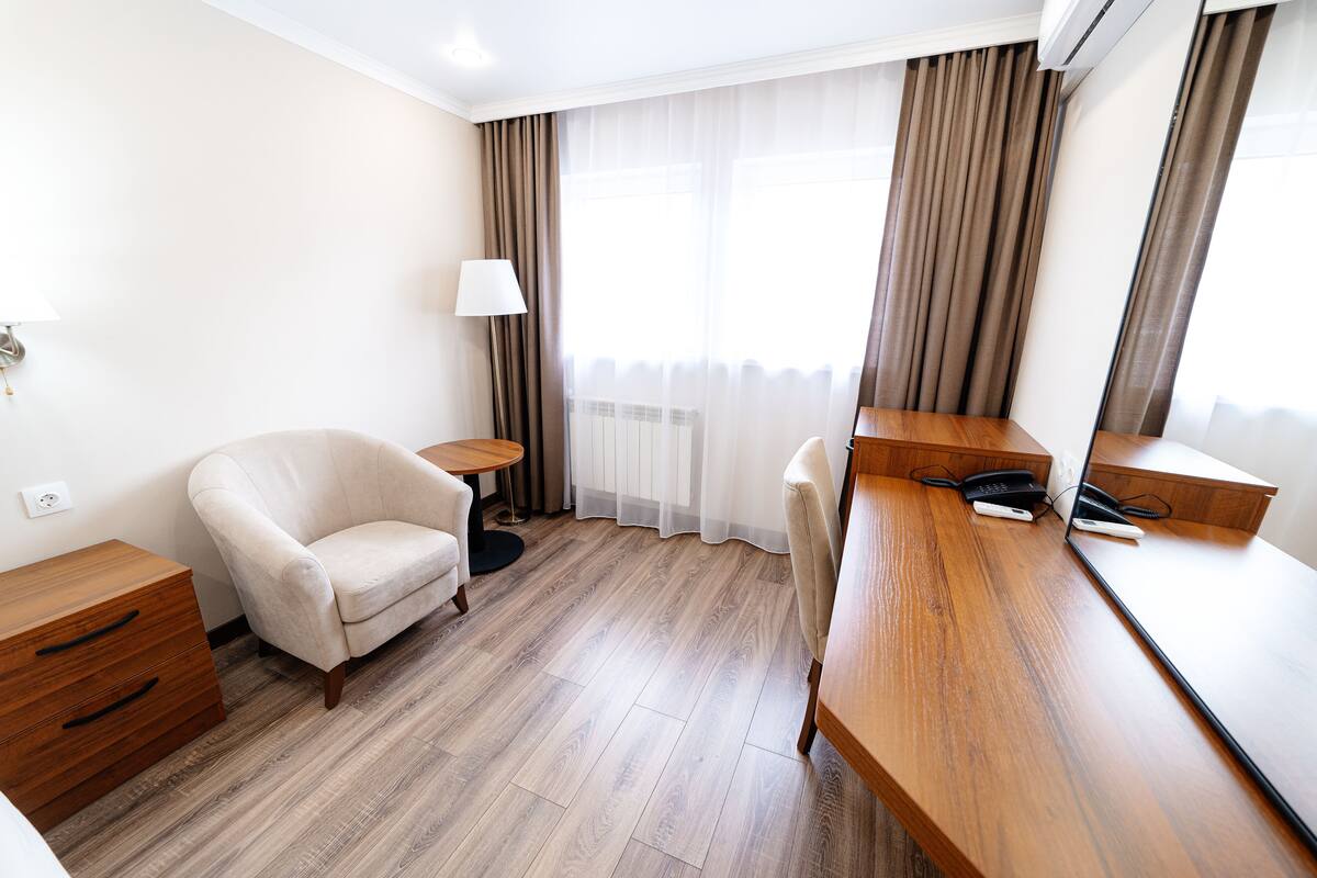 A hotel room with a wooden floor, a desk with a telephone, a chair, a bedside table, and a lamp.