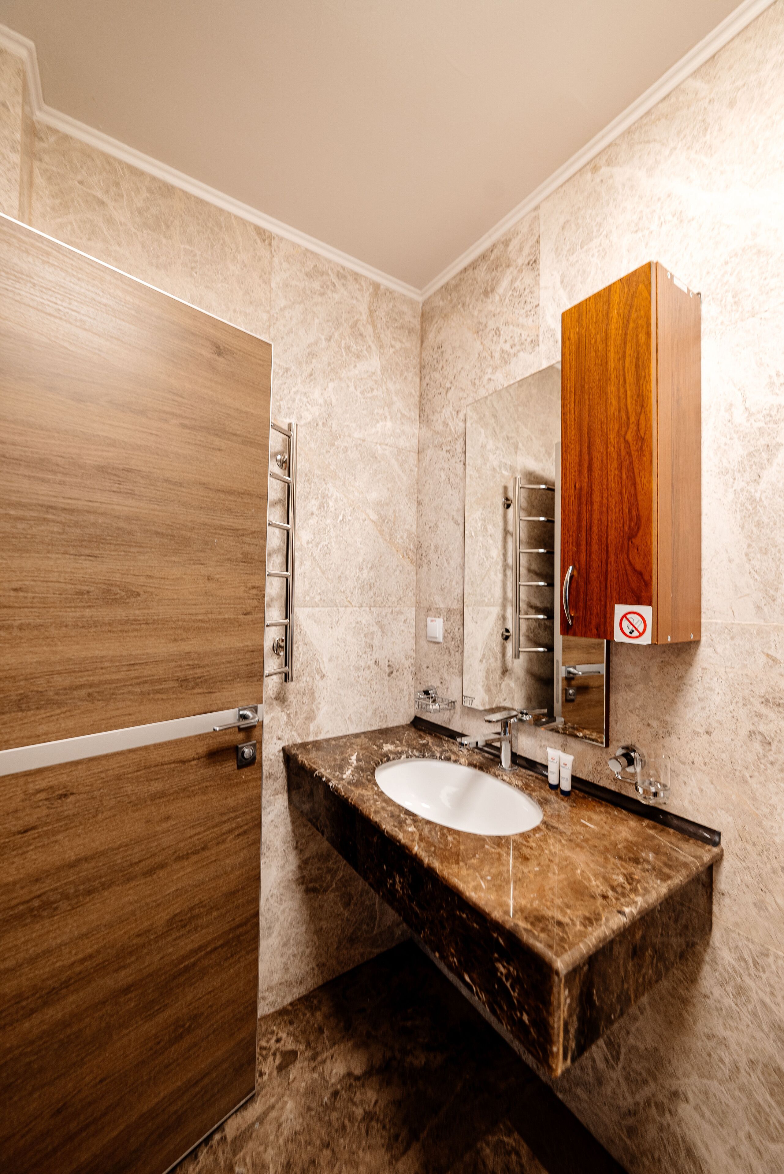 Un baño con puerta de madera, lavamanos de mármol y un mueble de madera sobre el espejo.