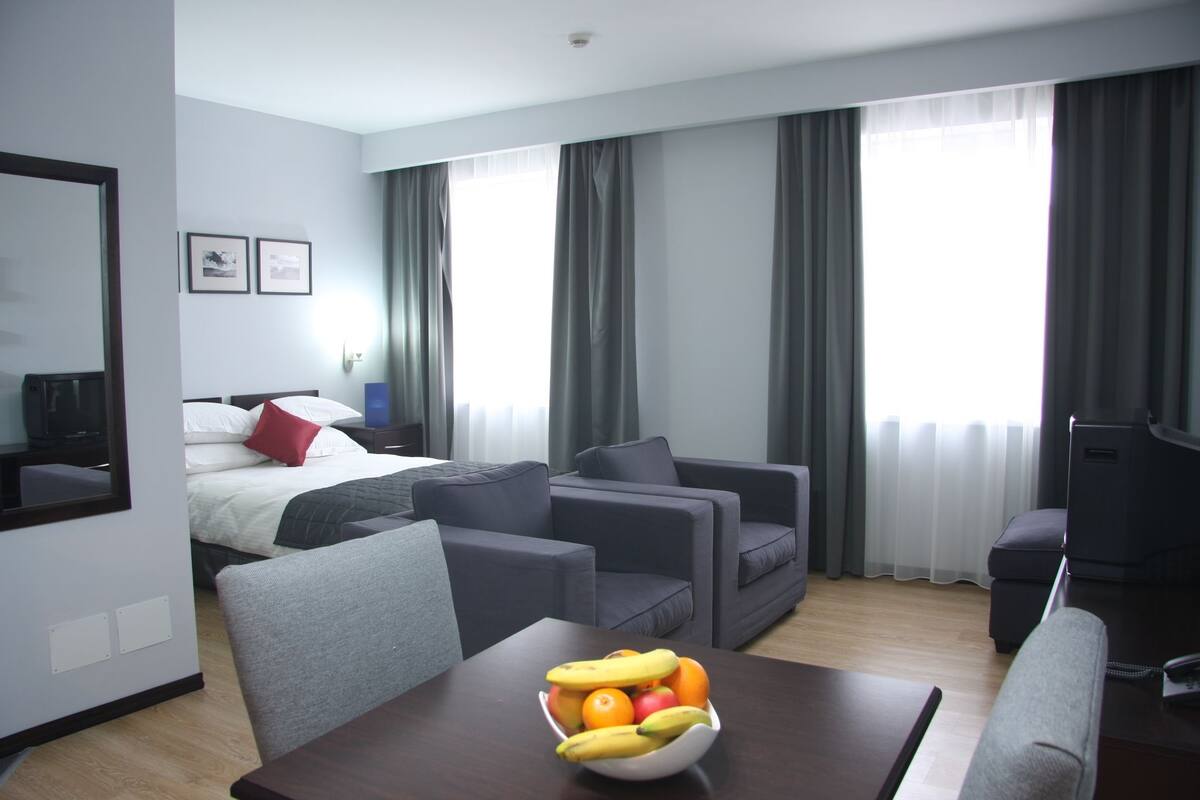 A hotel room with a bed, two sofas, a TV, a table with fruit, and large windows with curtains.