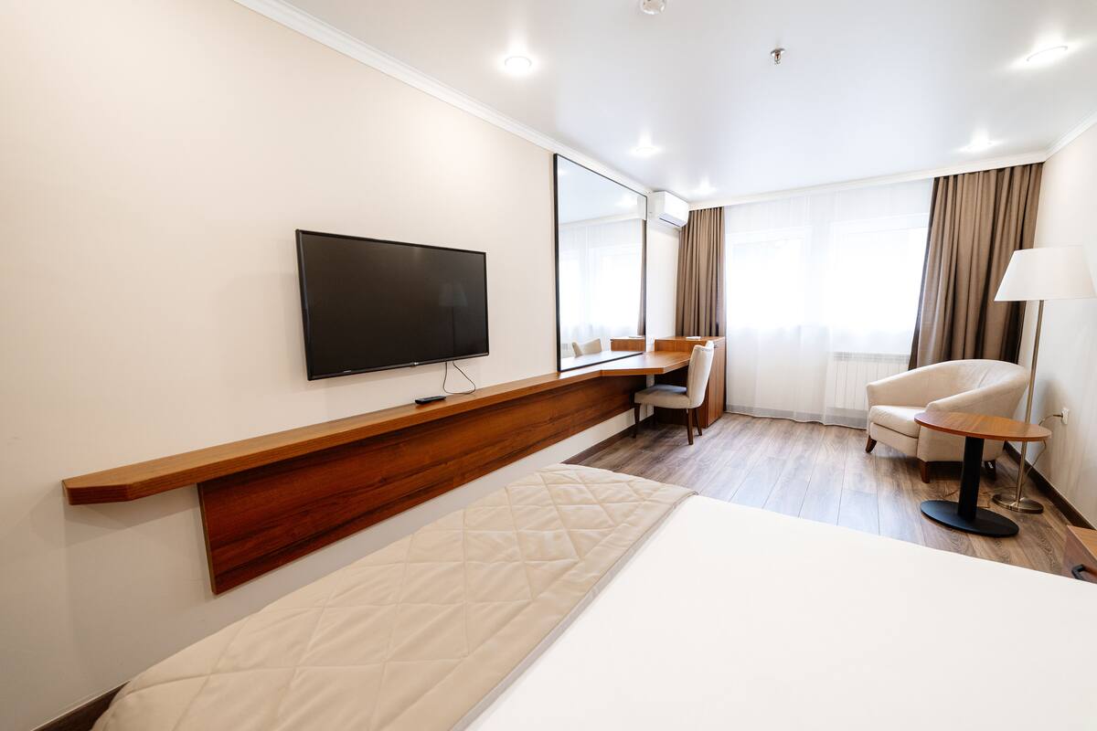 A hotel room with a large bed, a TV mounted on the wall, a desk with a chair, and a lamp.