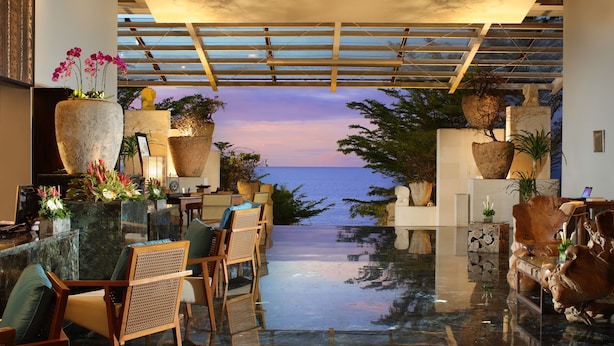 A modern lounge area with a view of the sea, featuring a glass floor, wooden furniture, and large planters. - Anantara Uluwatu Bali Resort (Pecatu)