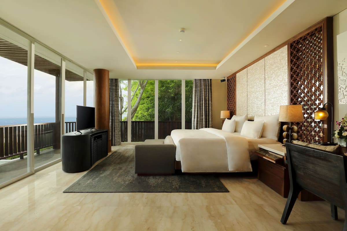 A modern hotel room with a large bed, a desk, a chair, a TV, and a balcony with a view of the sea.
