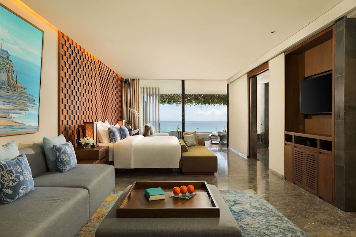 A modern hotel room with a large bed, a sofa, a coffee table with books and oranges, a TV, and a view of the sea.
