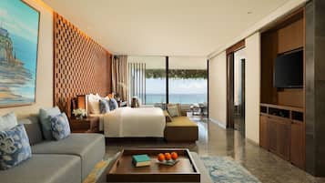 Quarto de hotel moderno com uma cama grande, um sofá, uma mesa de centro com livros e laranjas, uma televisão e vista para o mar.