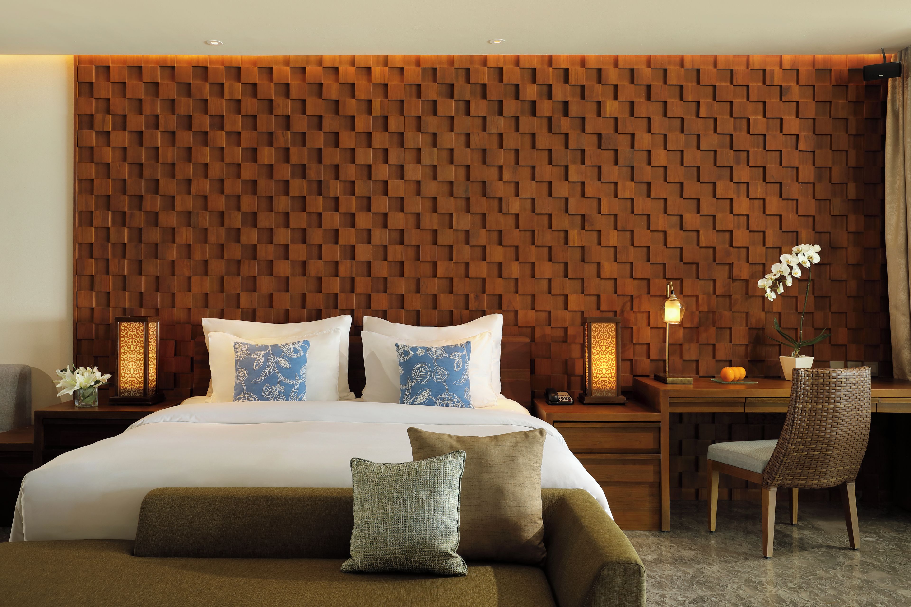 A hotel room with a wooden headboard, a bed with white bedding, two bedside tables with lamps, a chair, and a small table with a potted plant.