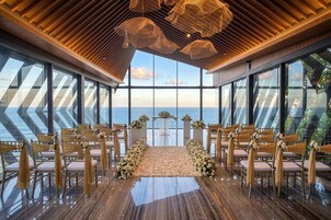 A wedding ceremony setup with a view of the sea, featuring a wooden floor, chairs with draped fabric, and floral arrangements.