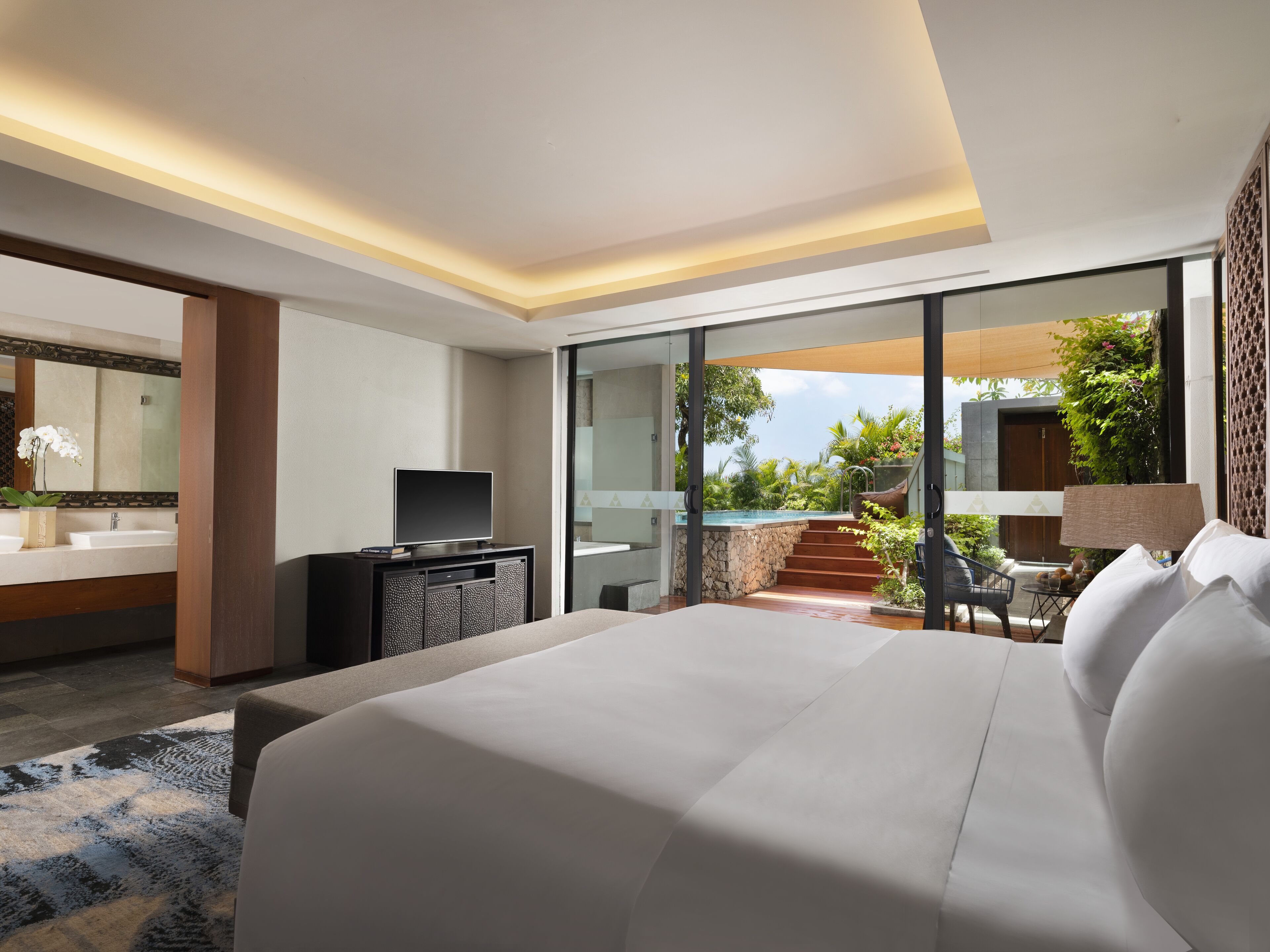 A modern hotel room with a large bed, a TV, a desk, and a view of a pool and garden.
