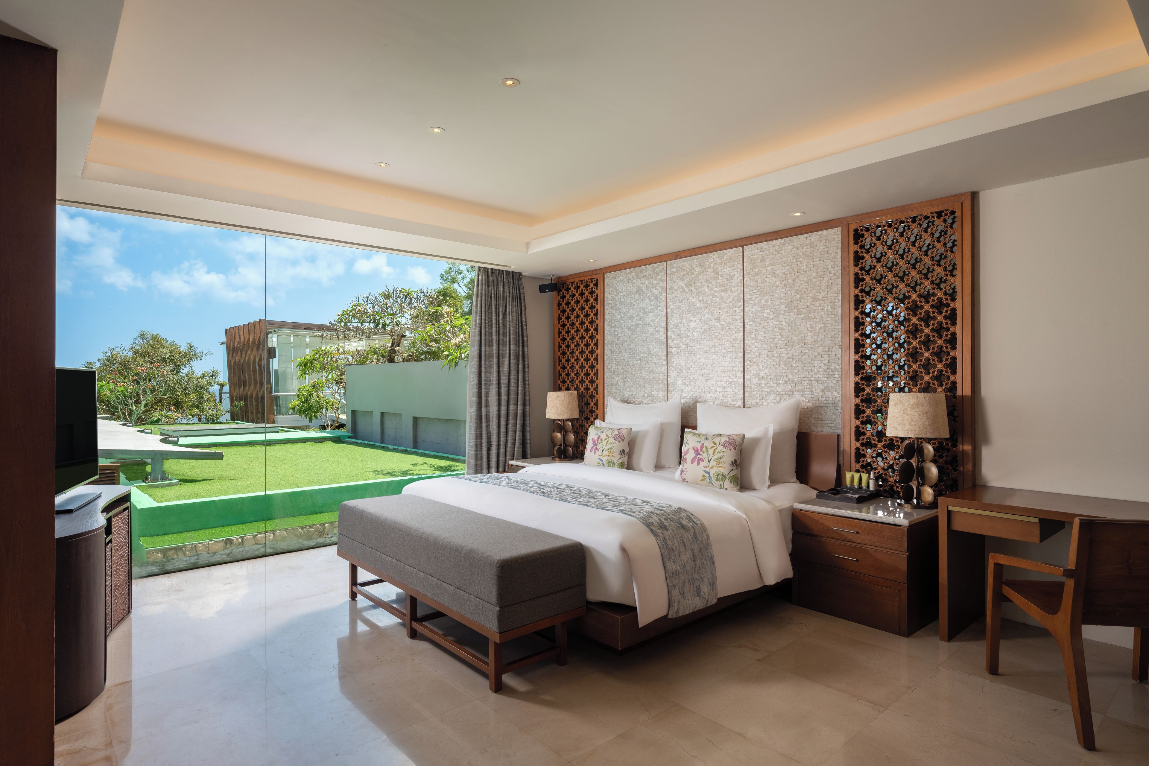 A modern bedroom with a large bed, a desk, and a view of a green lawn and building.
